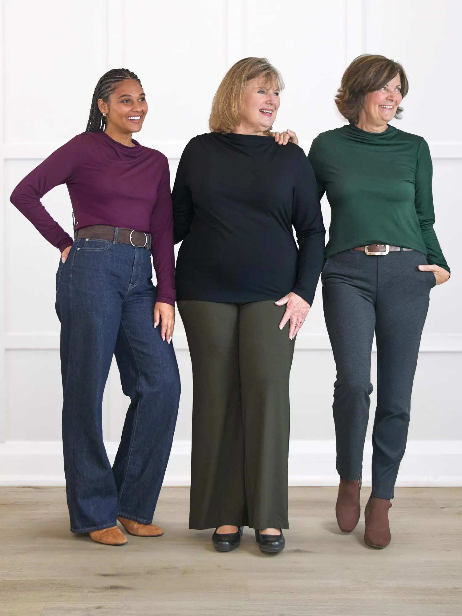 Three women standing together wearing different colored long-sleeve shirts and jeans against a white wall.
