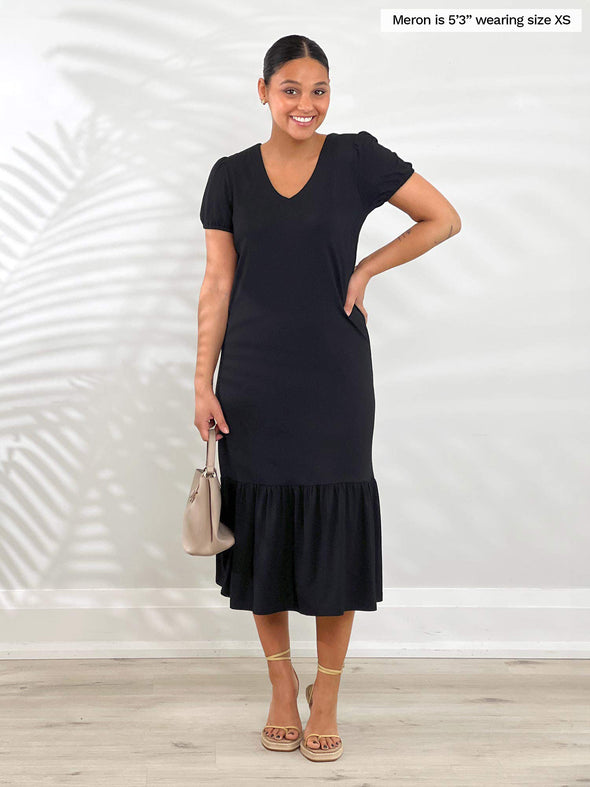 Miik model Meron (5'3", xsmall) smiling while standing in front of a white wall wearing Miik's Zilma reversible puff sleeve midi dress in black 