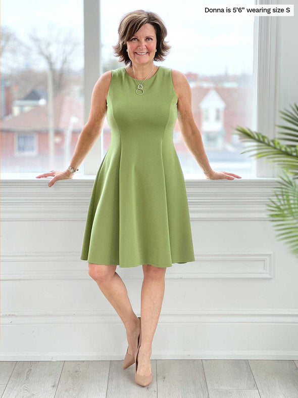 Miik founder Donna (five feet six, size small) smiling while standing in front of a window wearing Miik's Valerie sleeveless fit and flare dress in green moss