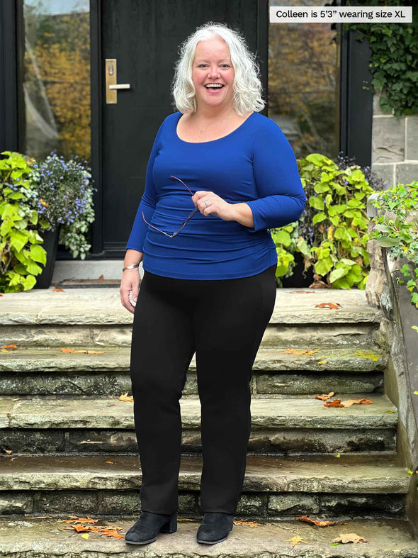 Miik model Colleen (5'3", xlarge) laughing while standing in front of a front door stairs wearing a black pant with Miik's Akari side ruched reversible top in blueberry 