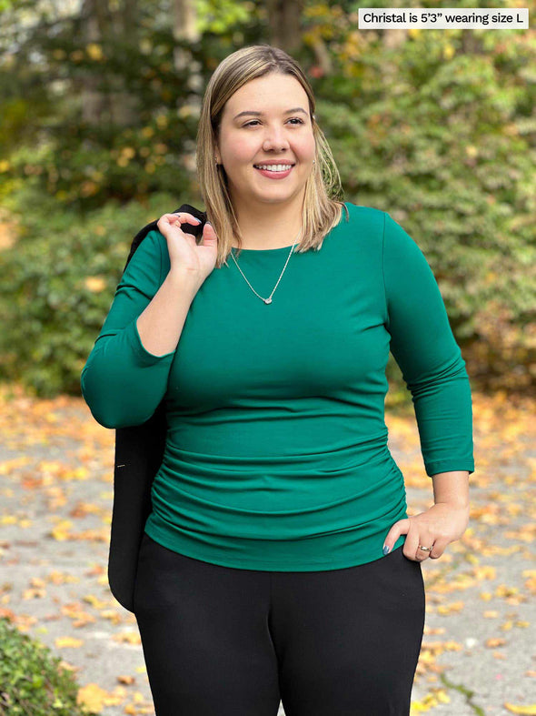 Miik model Christal (5'3", large) smiling while looking away wearing a black jogger with Miik's Akari side ruched reversible top in emerald green