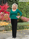Miik model Carolyn (5'10", large) smiling standing in front of a garden wearing Miik's Akari side ruched reversible top in emerald green with a black pant 