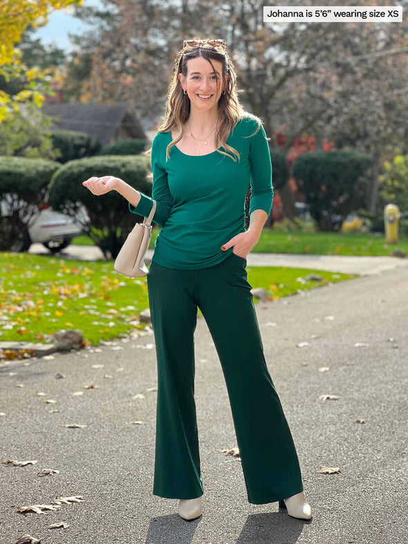 Miik model Jo (5'6", xsmall) smiling while standing in a side road wearing a pine green flare pant, Miik's Akari side ruched reversible top in emerald green and natural accessories 