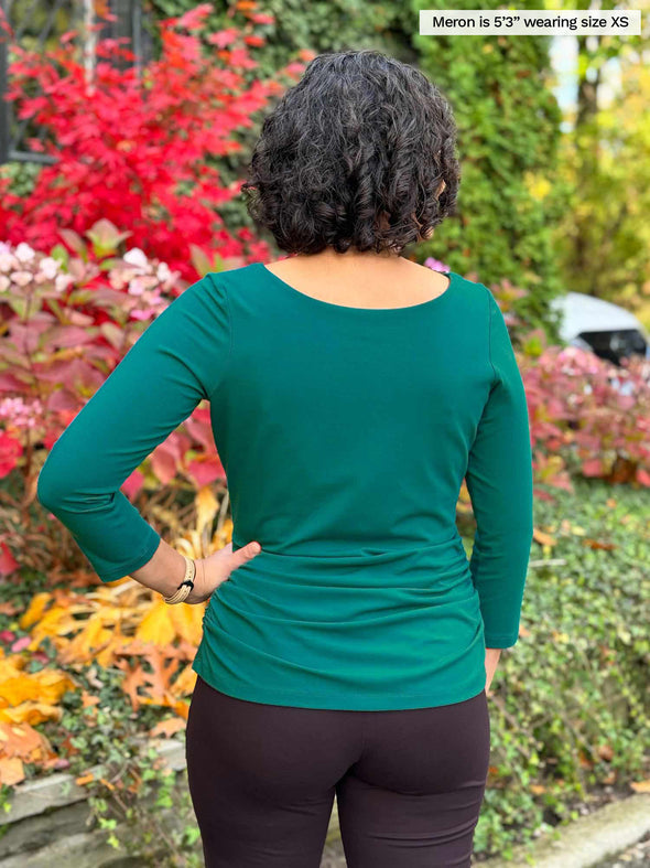 Miik model Meron (5’3”, xsmall) standing with her back towards the camera showing the back of Miik's Akari side ruched reversible top in emerald green