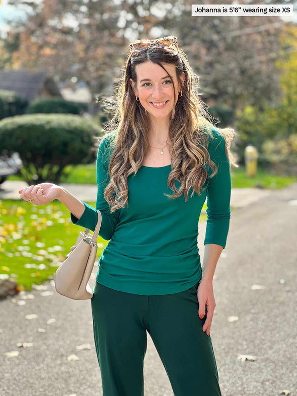 Miik model Jo (5'6", xsmall) smiling wearing Miik's Akari side ruched reversible top in emerald green along with a flare pant in pine green 