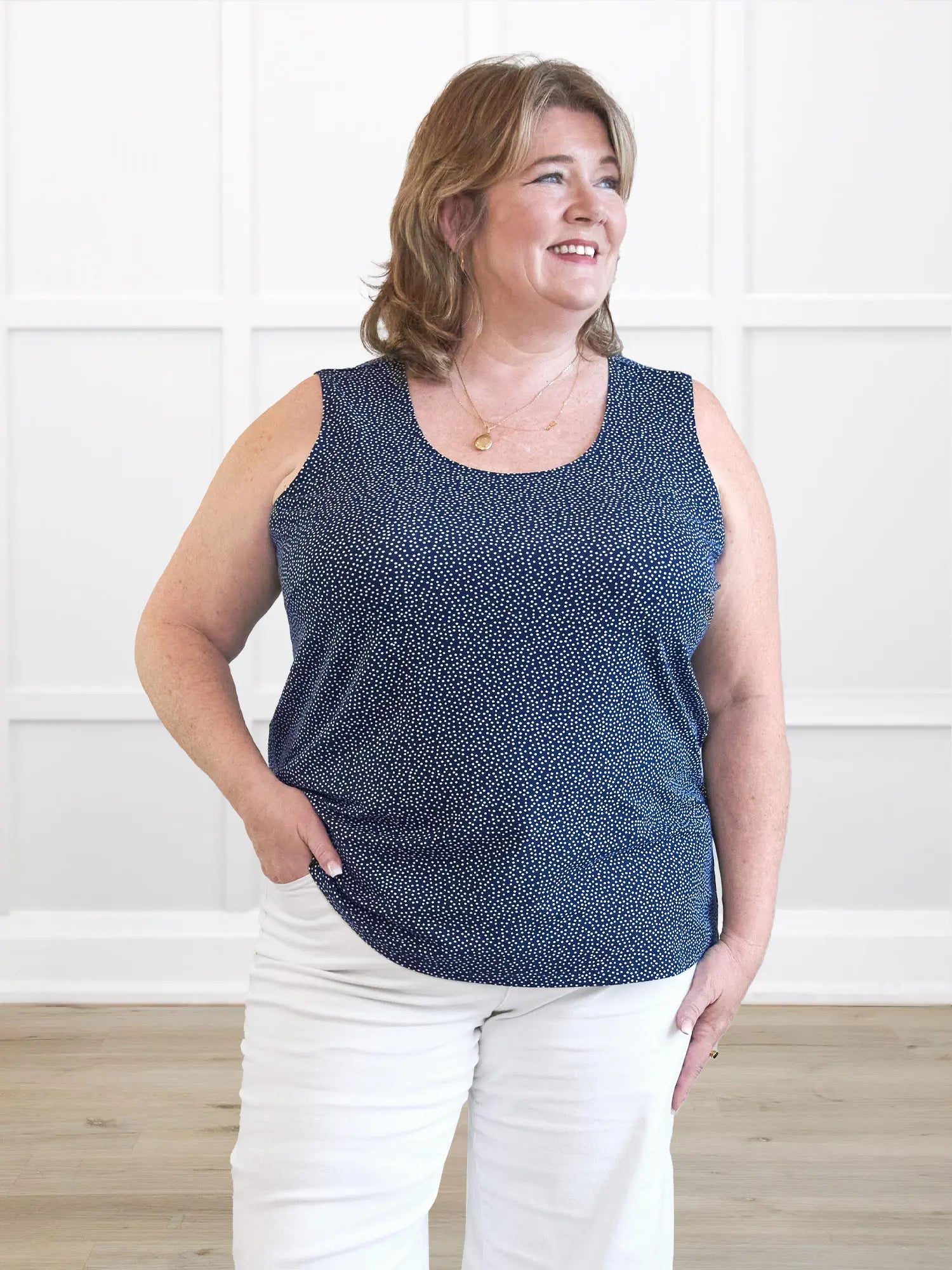 Woman wearing a blue patterned sleeveless top and white pants against a white wall.