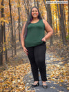 Miik model Sureka (5'9", size 2X) smiling while standing in nature (fall leafs) wearing Miik's Alanis relaxed tank top in pine green along with a dress pant in black 