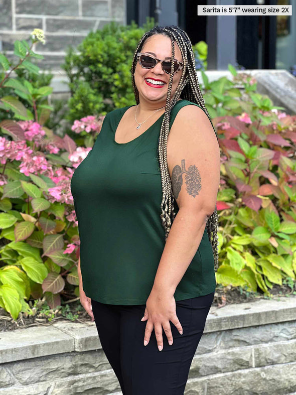 Miik model Sarita (5'7", 2x) smiling while standing sideway wearing Miik's Alanis relaxed tank top in pine green with a black pant and sunglasses