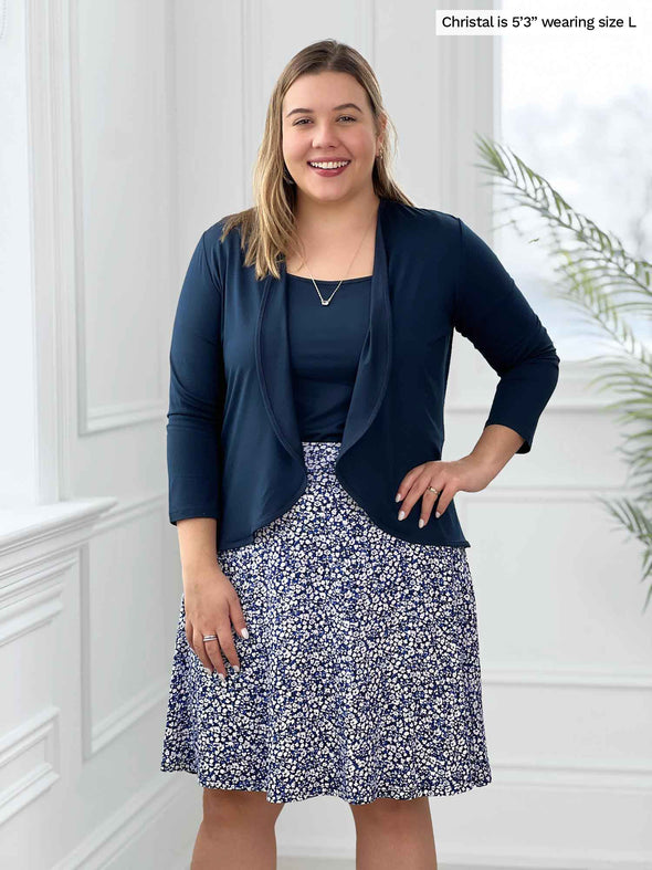 Miik model Christal (5'3", large) smiling while standing in front of a white wall wearing a tank top and a cardigan in navy with Miik's Alara pocket swing skirt in baby's breath print 