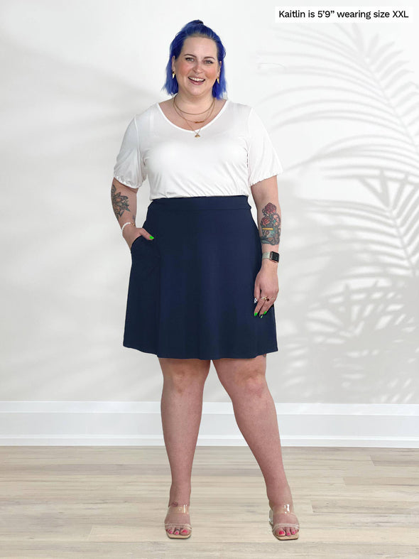 Miik model Kaitlin (5'9, xxlarge) smiling white standing in front of a white wall wearing Miik's Alara pocket swing skirt in navy with a white tee