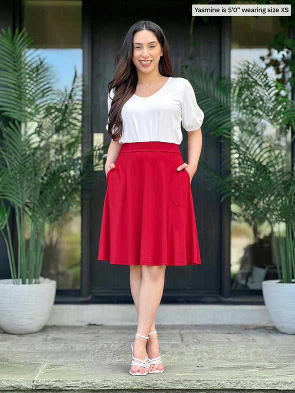 Miik model Yasmine (5'0", xsmall, petite) smiling with hands on pockets wearing a white tee top along with Miik's Alara pocket swing skirt in poppy red 