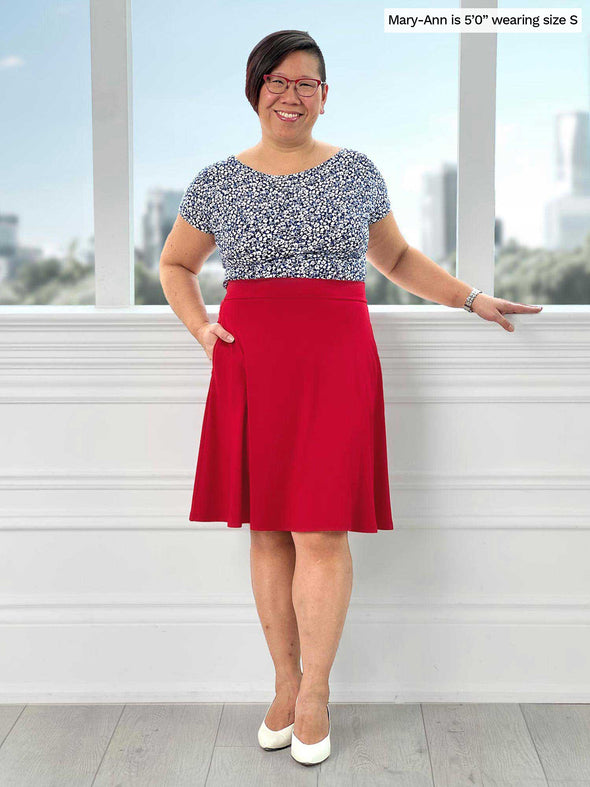 Miik model Mary-Ann (5'0, small, petitte) smiling while standing in front of a window wearing Miik's Alara pocket swing skirt in poppy red with a printed tee 