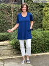 Miik founder Donna (5'6", small) smiling with one hand on pockets wearing Miik's Alma half sleeve pocket tunic in ink blue with a white jeans 