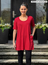 Miik model Meron (5'3", xsmall) smiling while standing in a door entry wearing Miik's Alma half sleeve pocket tunic in poppy red with black legging 