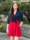 Miik model Yasmine (5'0", xsmall, petite) smiling and looking down wearing Miik's Alma half sleeve pocket tunic in poppy red as a dress with a cardigan in navy tie