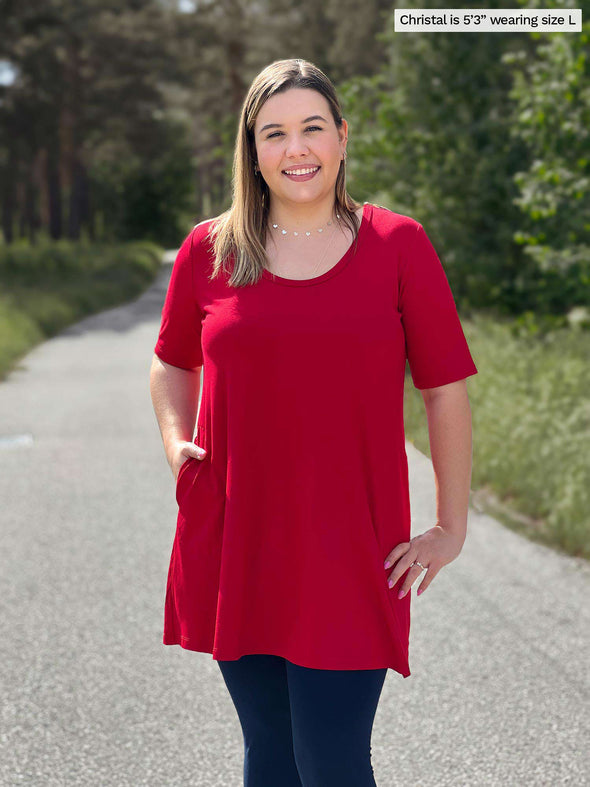 Miik model Christal (5'3", large) smiling wearing Miik's Alma half sleeve pocket tunic in poppy red with a navy legging 