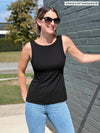 Miik model Johanna (5'6", xsmall) standing next to a brick wall smiling wearing Miik's Amina reversible shelf bra tank in black reversed, jeans and sunglasses  