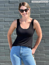 Miik model Johanna (5'6", xsmall) standing in front of a brick wall smiling wearing Miik's Amina reversible shelf bra tank in black with jeans and sunglasses 