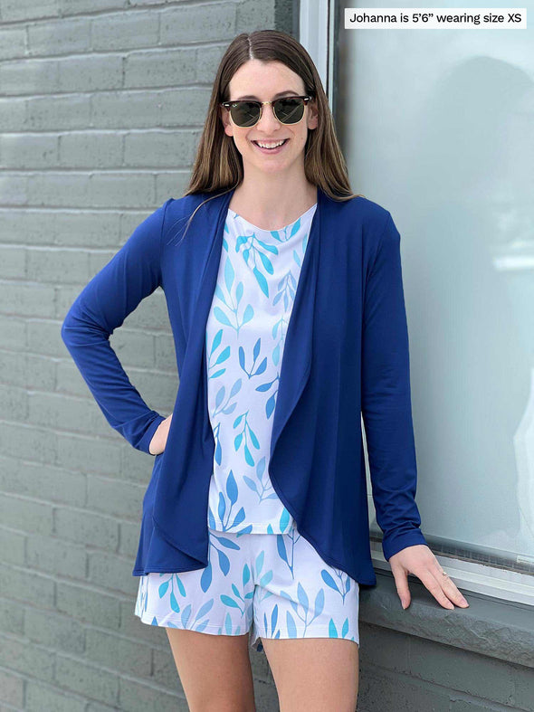 Miik model Johanna (five feet five, xsmall) smiling and standing next to a brick wall/window wearing Miik's Amita PJ lounge set in ocean leaf print with a blue cardigan