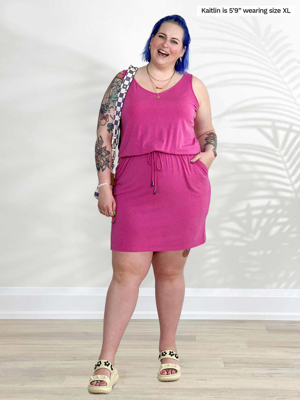 Miik model Kaitlin (5'9", xlarge) smiling while standing in front of a white wall wearing Miik's Amy drawstring pocket dress in pink 