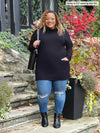 Miik model Carley (5'2", xxlarge) smiling while standing in front of a door way wearing Miik's 		Ashanti long sleeve cowl neck pocket tunic in black, ripped jeans and black boots and shoulder tote bag