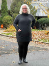 Miik model Colleen (5'3", xlarge) smiling with hands on pockets wearing Miik's 		Ashanti long sleeve cowl neck pocket tunic in charcoal, black leggings and black boots 