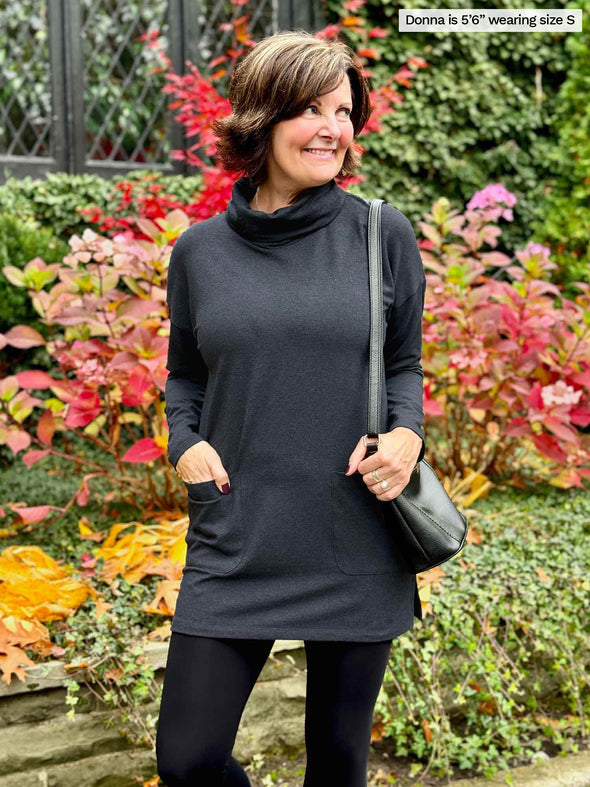 Miik founder Donna (5'6", small) smiling and looking away wearing Miik's 		Ashanti long sleeve cowl neck pocket tunic in charcoal with black leggings 
