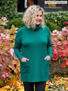 Miik model Carolyn (5'10", large) smiling and looking away with hands on pockets wearing Miik's 		Ashanti long sleeve cowl neck pocket tunic in jade melange and charcoal leggings 