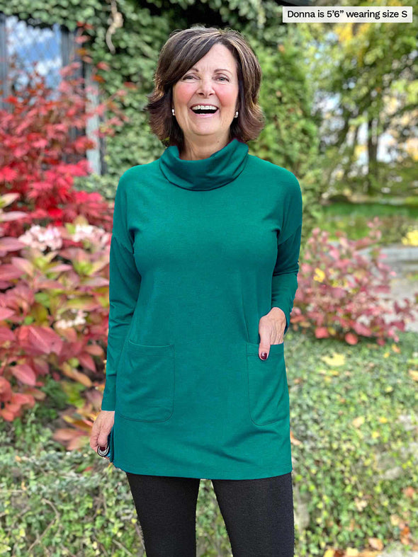 Miik founder Donna (5'6", small) smiling with one hand on pockets wearing Miik's 		Ashanti long sleeve cowl neck pocket tunic in jade melange and leggings 