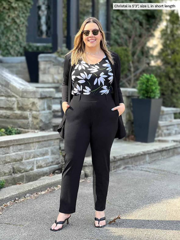 Miik model Yasmine (5'0", xsmall, petite) looking down wearing Miik's Asia mid-rise slim pant short length in black with a tank top in camel melange tucked in and high heels