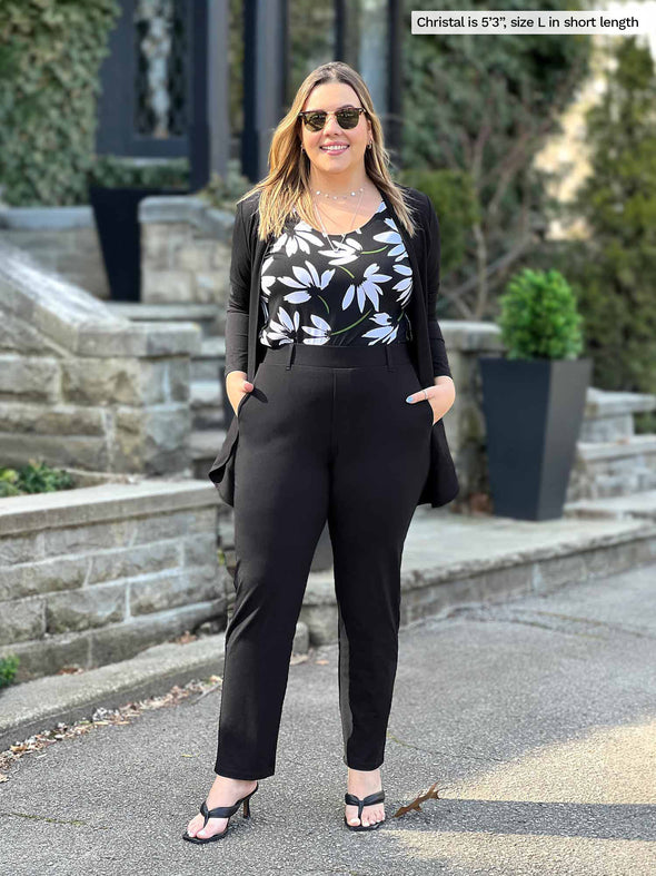 Miik model Christal (5'3", large) smiling wearing Miik's Asia mid-rise slim pant black in short length with a white lily tank top and a black cardigan