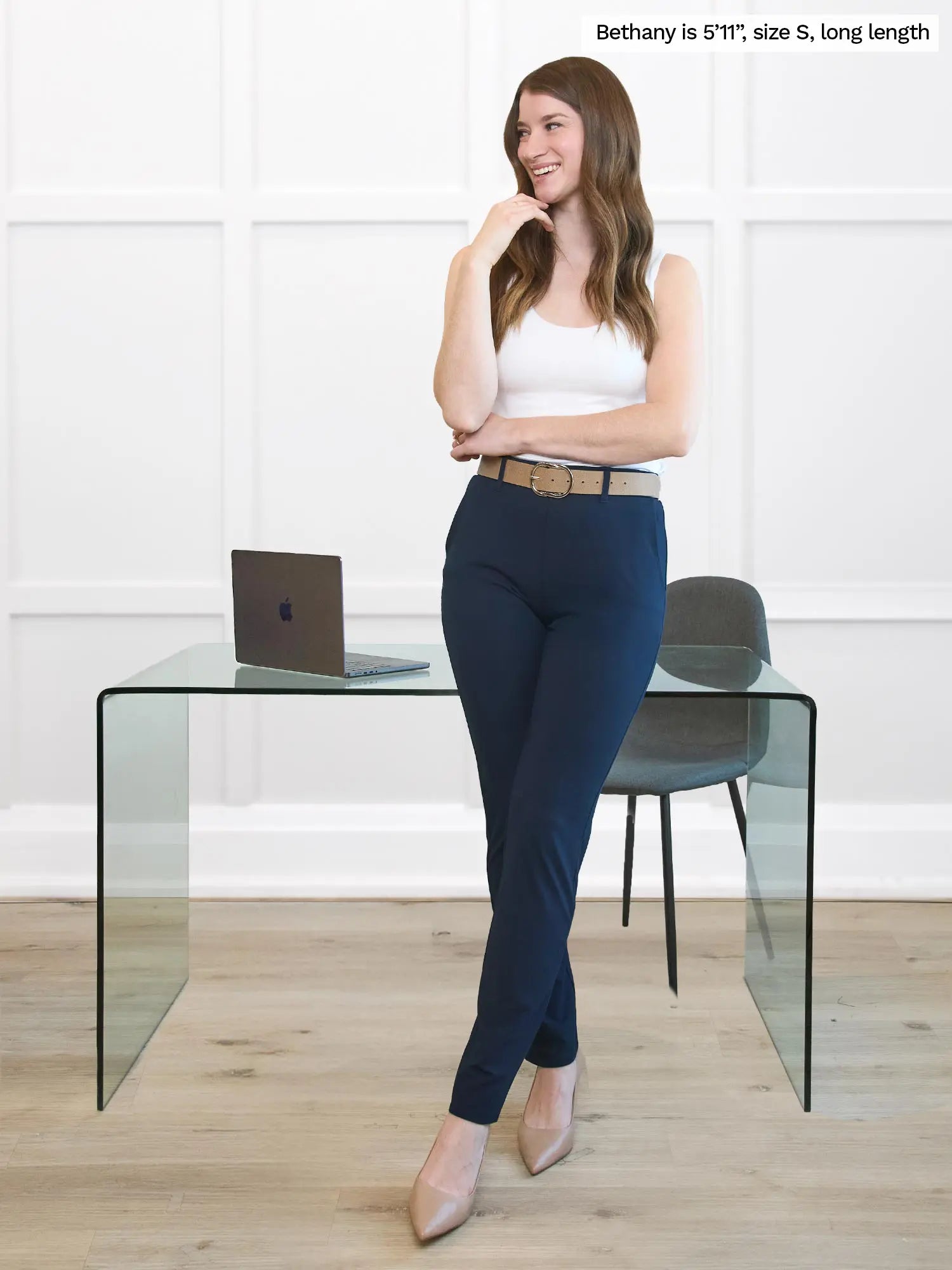 Woman wearing navy pants standing next to a glass table with a laptop. 