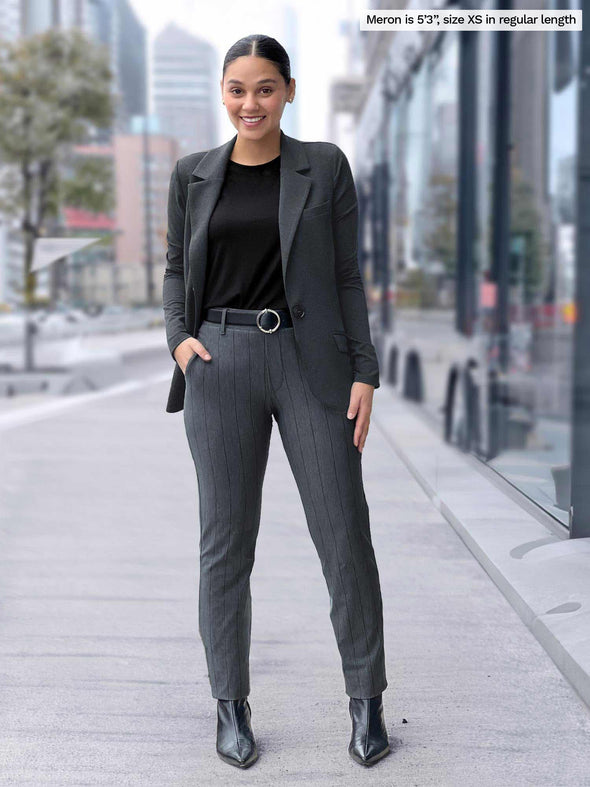 Miik model Meron (5'3", xsmall) smiling wearing Miik's Asia mid-rise slim pant in granite wide pinstripe with a charcoal blazer and a black top, belt and boots 