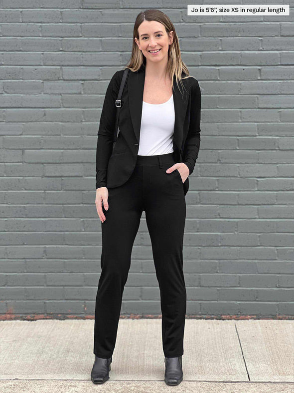 Woman standing in front of a brick wall wearing Miik's Asia mid-rise slim pant in black with a matching colour blazer and a white tank