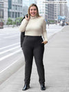 Miik model Christal (5'3", large) smiling wearing Miik's Asia mid-rise slim pant in charcoal wide pinstripe with a turtleneck top in oatmeal melange, black boots and a charcoal blazer over her shoulders 