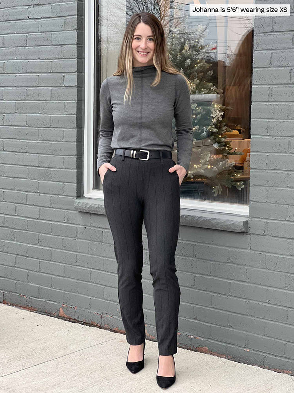 Woman smiling standing in front of a window wearing Miik's Asia mid-rise slim pant in charcoal wide pinstripe with a fleece top in charcoal and a black belt