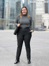 Woman standing in front of a window looking away wearing Miik's Asia mid-rise slim pant in charcoal wide pinstripe with a charcoal fleece top and a black belt