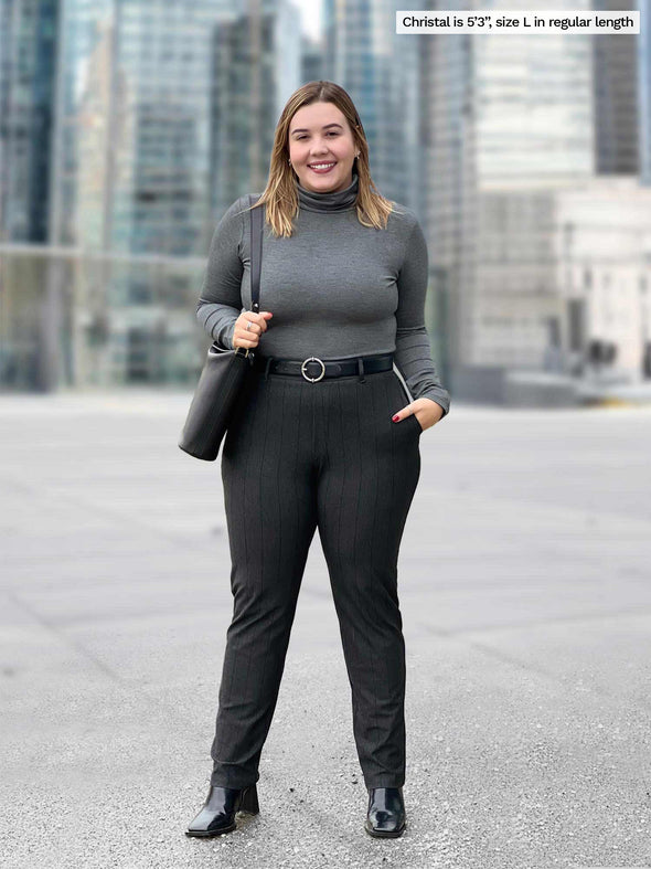 Woman standing in front of a window looking away wearing Miik's Asia mid-rise slim pant in charcoal wide pinstripe with a charcoal fleece top and a black belt