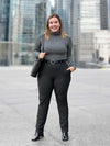 Miik model Christal (5'3", large) smiling wearing Miik's Asia mid-rise slim pant in charcoal wide pinstripe with a turtleneck top in granite melange, black boots, belt and tote bag