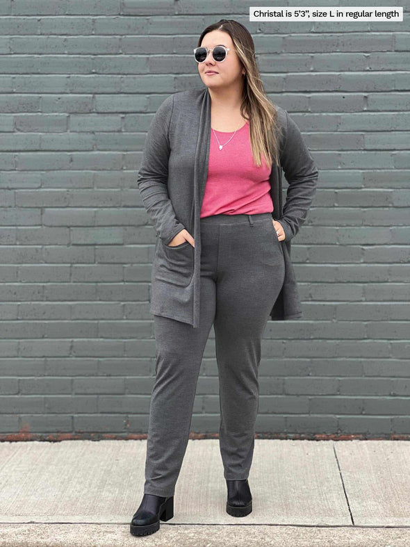 Miik model Christal (5'3", large) looking away while standing in front of a brick wall wearing Miik's Asia mid-rise slim pant in granite melange with a long fleece cardigan in the same colour and a pink tank top 