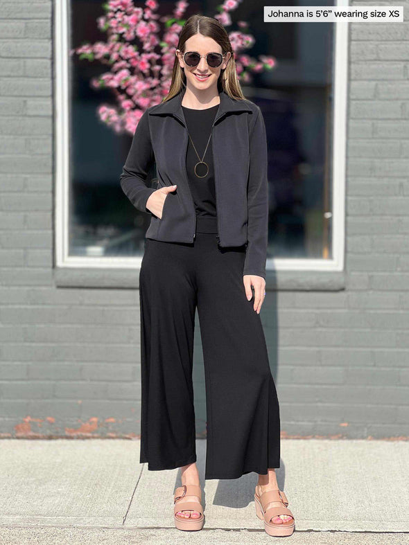 Miik model Johanna (five feet six, size xsmall) smiling while standing in front of a building wearing an all black outfit: wide leg pants and a basic tank with Miik's Ayesha zipped jacket in graphite