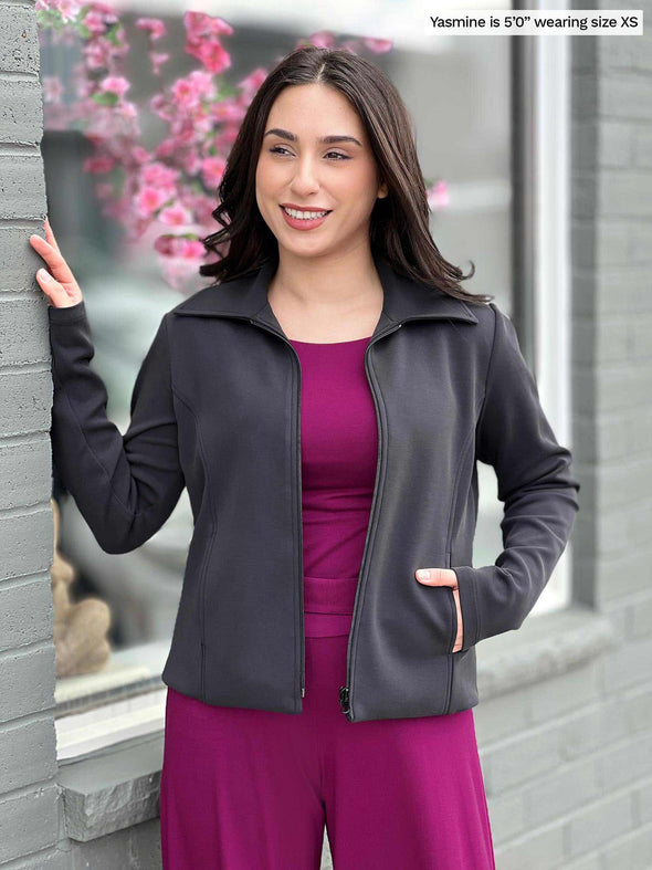 Miik model Yasmine (five feet tall, size xsmall, petite) standing next to a brick wall and smiling wearing Miik's Ayesha zipped jacket in graphite along with a top and wide leg pant in ruby