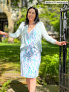 Woman standing in nature next to a fence smiling while wearing Miik's benton wrap dress in blue leaf pattern along with short blue cardigan
