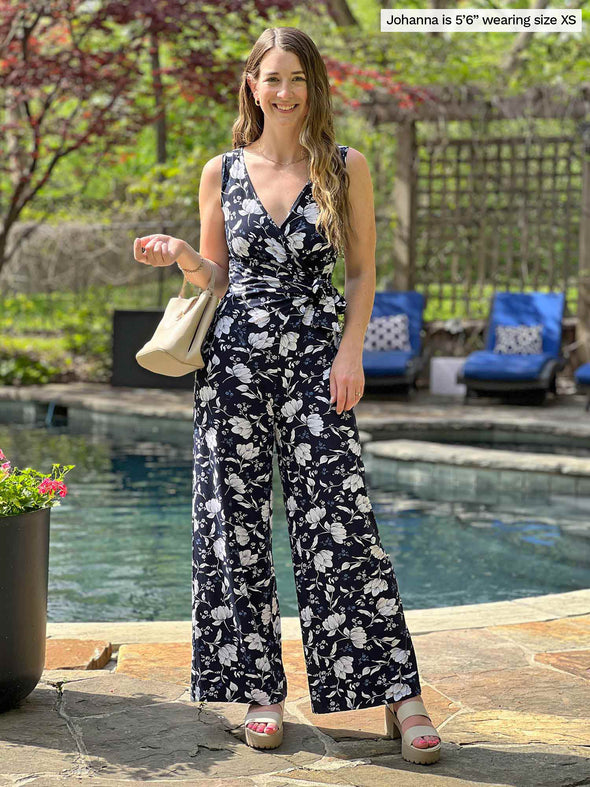 Miik model Johanna (five feet six, xmall) smiling in front of a pool holding a purse wearing Miik's Blakely faux-wrap dressy jumpsuit in blossom print