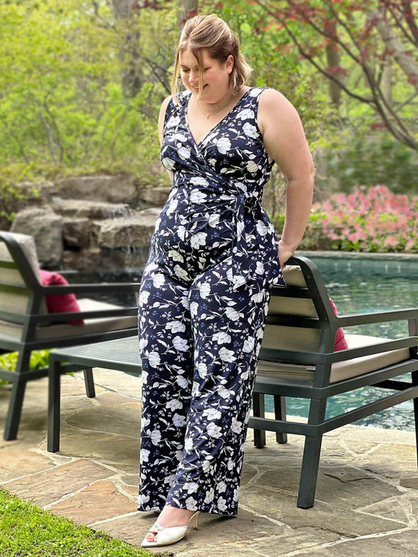 Miik model Bri (five feet five, xlarge) leaning agains to a chair in front of a pool wearing Miik's Blakely faux-wrap dressy jumpsuit in blossom print