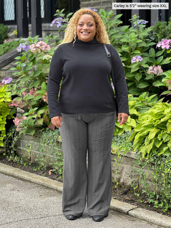 Miik model Carley (five feet two, xxlarge) smiling while standing next to a garden wearing Miik's Breda funnel neck long sleeve top in graphite with a pinstriped pant in graphite melange 