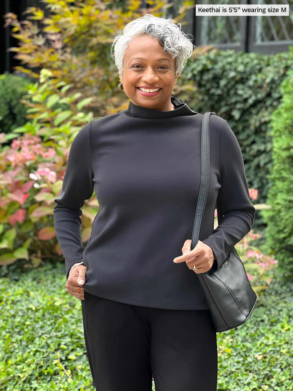 Miik model Keethai (five feet five, medium) standing in front of a garden smiling wearing Miik's Breda funnel neck long sleeve top in graphite with a black pant and a shoulder bag