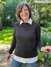 Miik founder Donna (five feet six, small) smiling while standing in a backyard wearing a collared white shirt under Miik's Breda funnel neck long sleeve top in graphite and jeans 