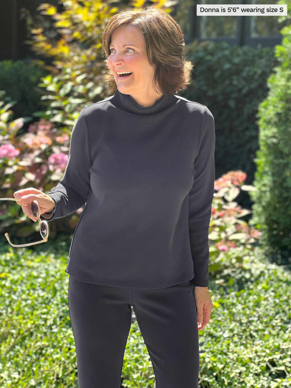 Miik founder Donna (five feet six, small) smiling while looking away wearing Miik's Breda funnel neck long sleeve top in graphite along with a lounge pant in the same colour 