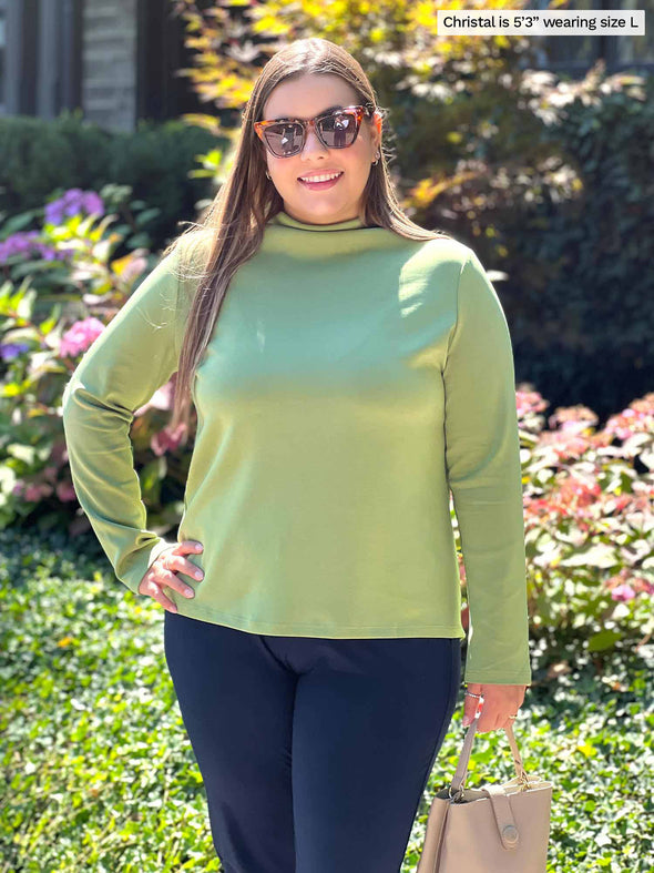 Miik model Christal (five feet three, large) smiling while wearing Miik's Breda funnel neck long sleeve top in moss with a navy pant  
